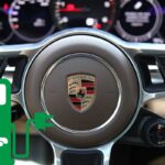 Il logo Porsche sul volante e in basso a sinistra il disegno di una colonnina per auto elettriche